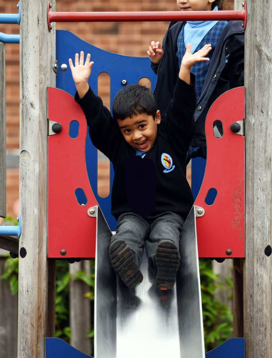 A boy slides down the slide in the Nursery playground with his arms in the air.