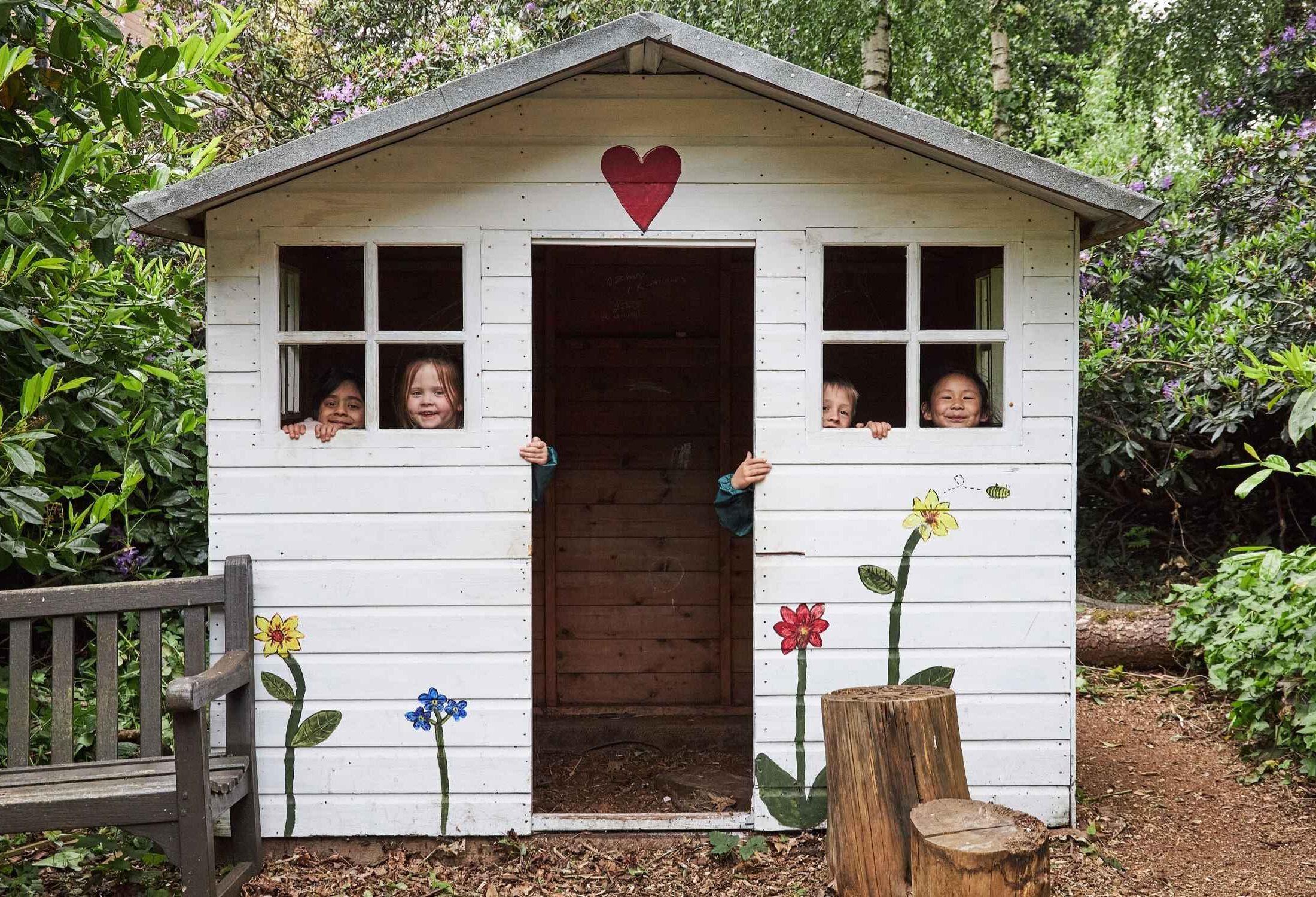 Four children looking through the windows of the forest cottage in Forest School.