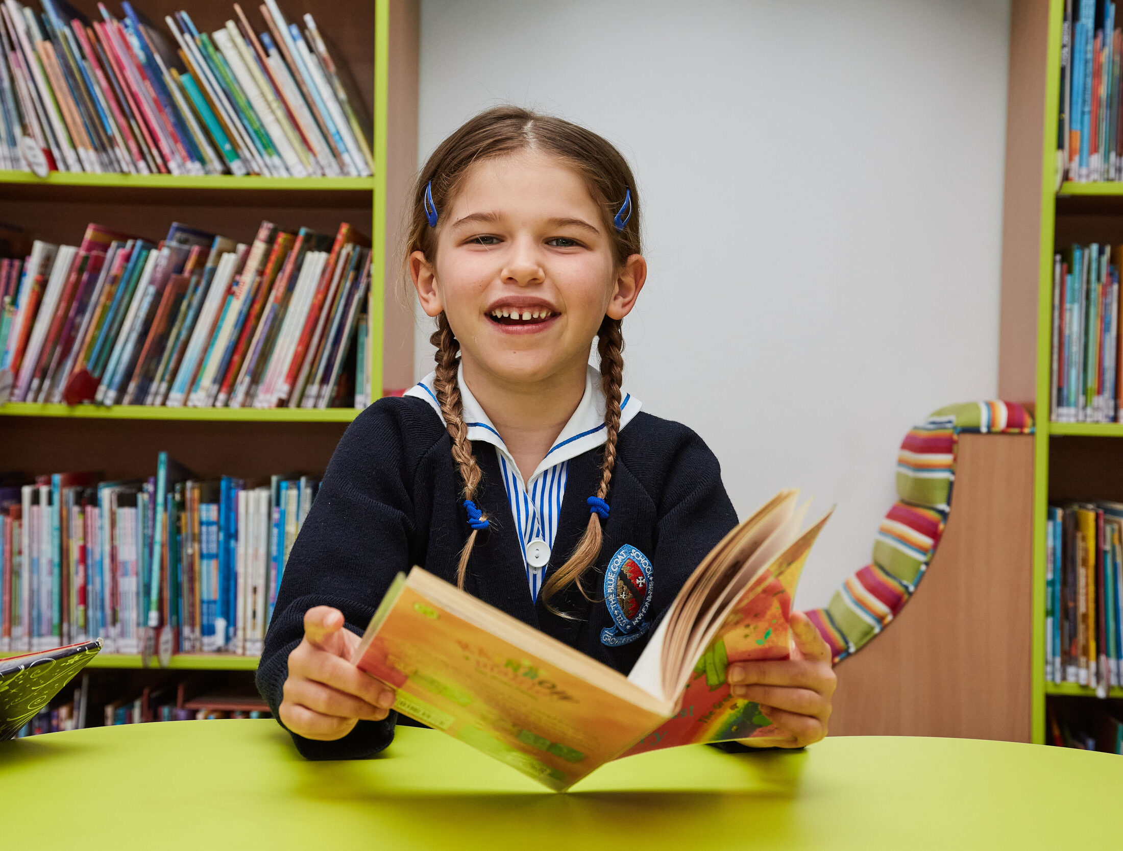 A girl sat in the Pre-Prep Library, smiling at the camera with an open book in her hand.