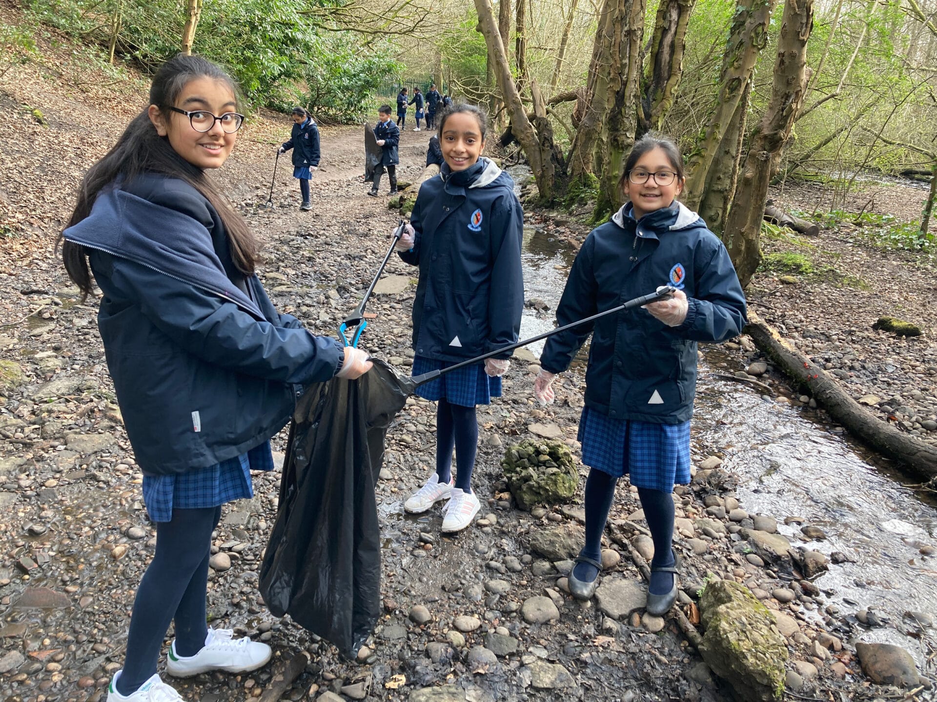 Three girls in Blue Coat School uniform using a litter picker in a woodland area during a community litter picking project.