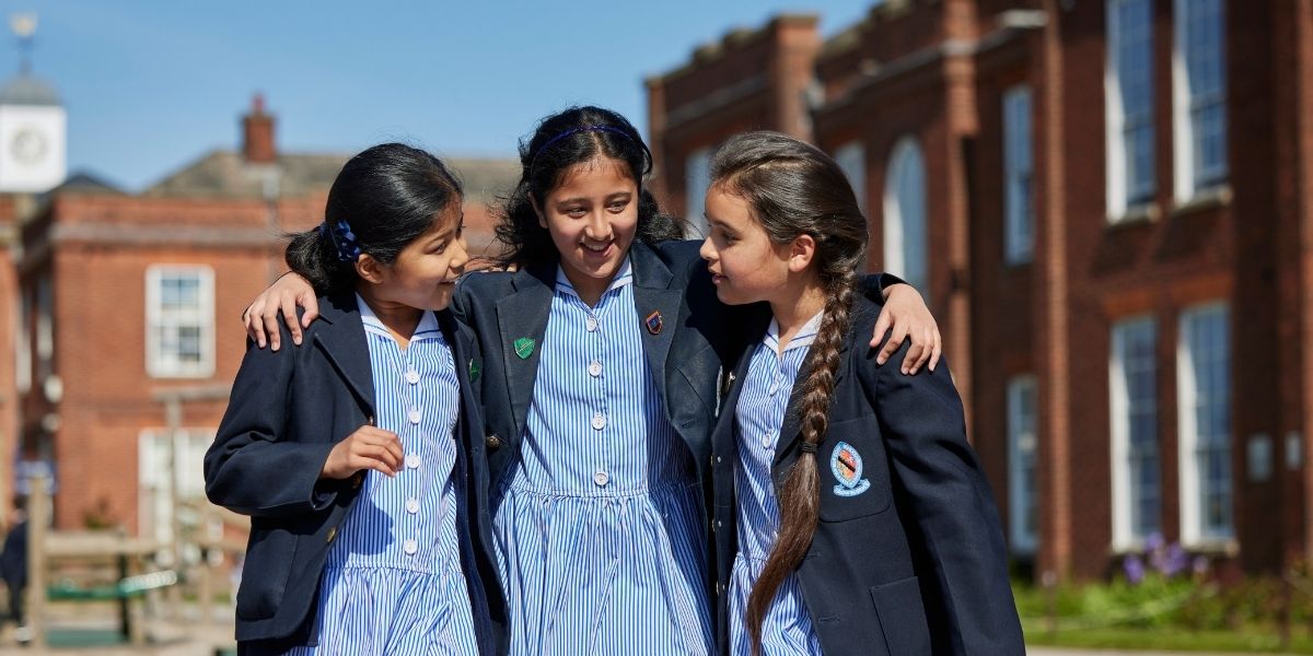 Three girls walk in the playground with their arms around each other, chatting and laughing.