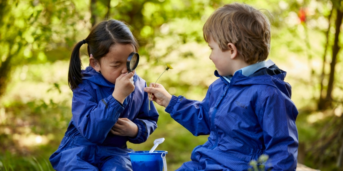 Two pupils sit on a log in Forest School looking through a magnifying glass at a flower.