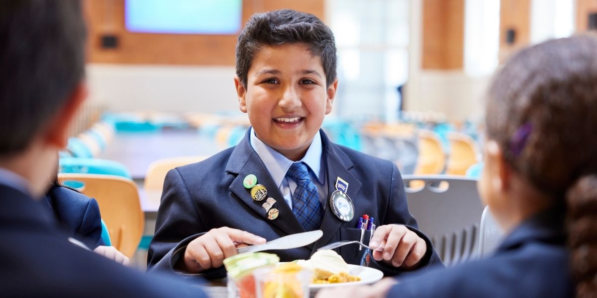 A boy is smiling at the camera as he eats his lunch in The Blue Coat School Dining Hall.