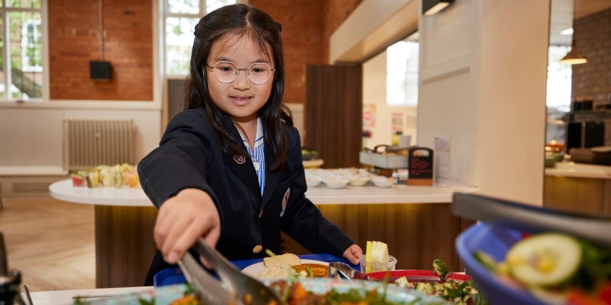 A pupil in Blue Coat uniform is using tongs to put some sweet potato salad on her plate in the Dining Hall.