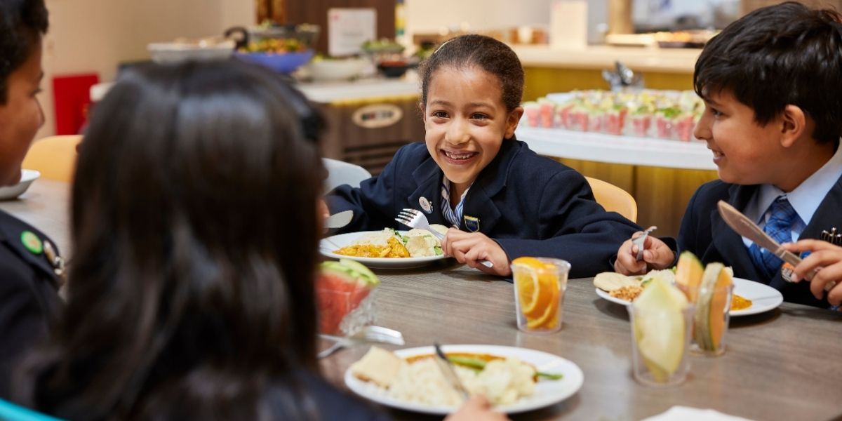 A group of pupils at The Blue Coat School Birmingham are sat at a lunch table in the Dining Hall eating curry and smiling at each other.