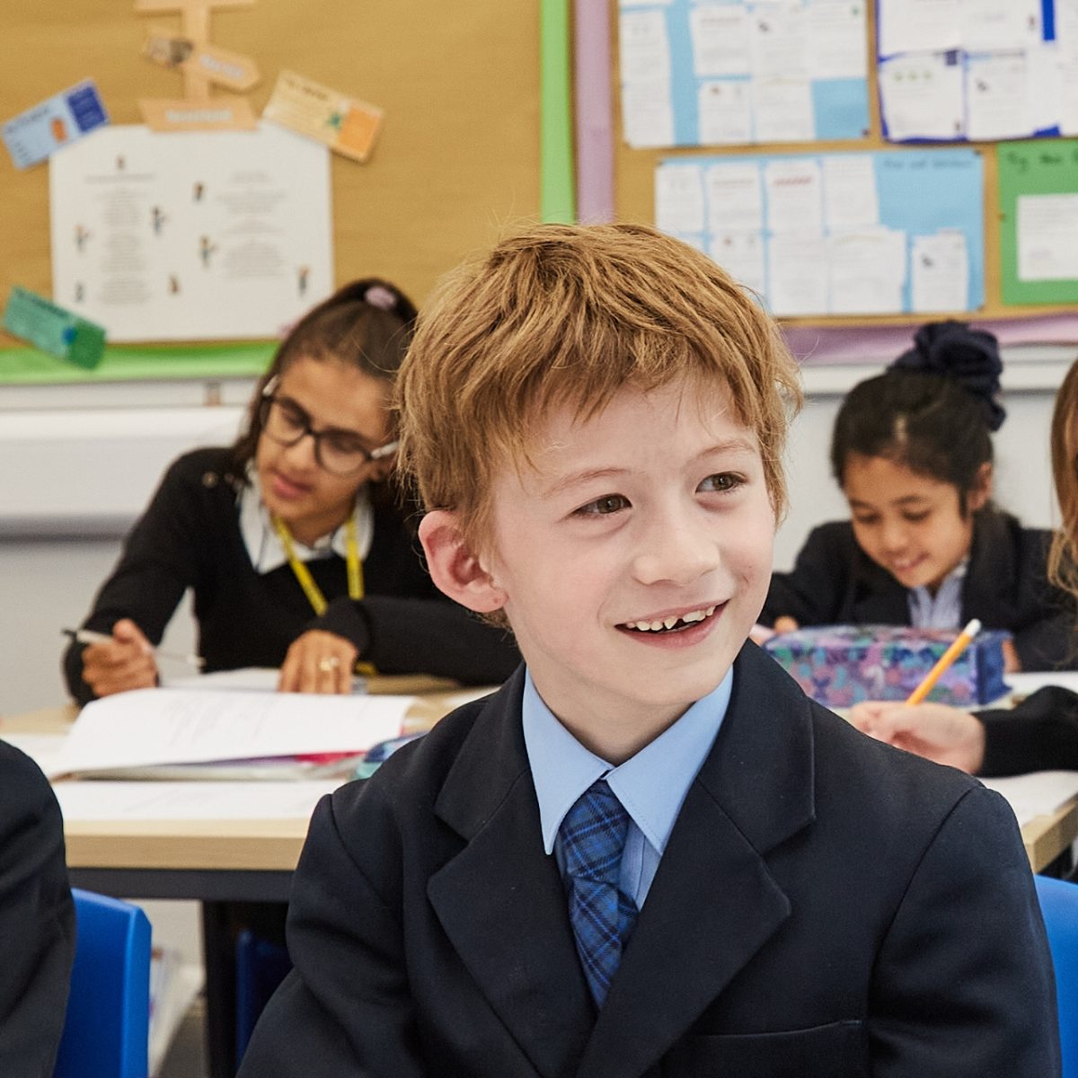 A boy smiles over at the teacher in the classroom with other children working in the background.